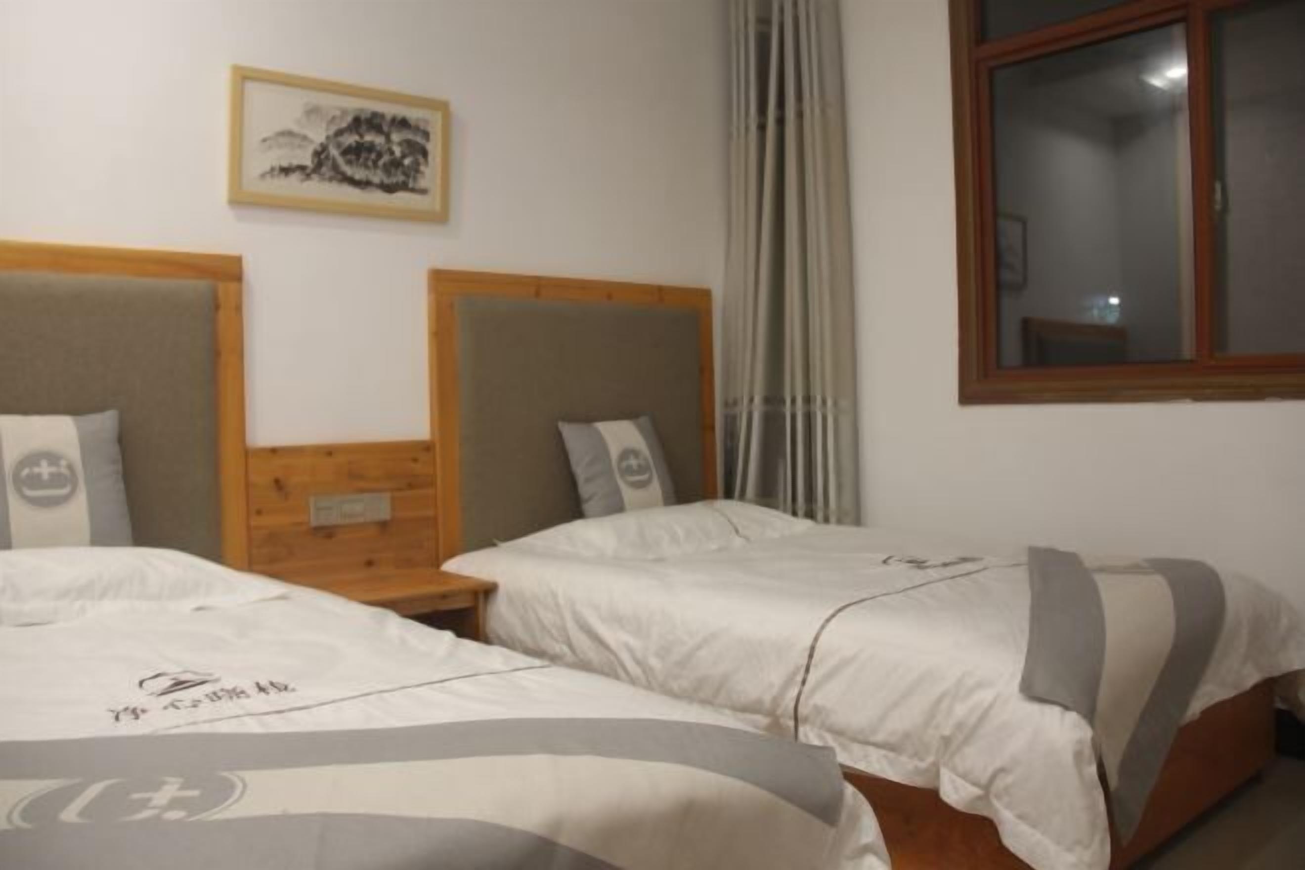 Habitación de hotel con dos camas, cabecero de madera, mesita de noche, un cuadro enmarcado en la pared y una ventana con cortinas.