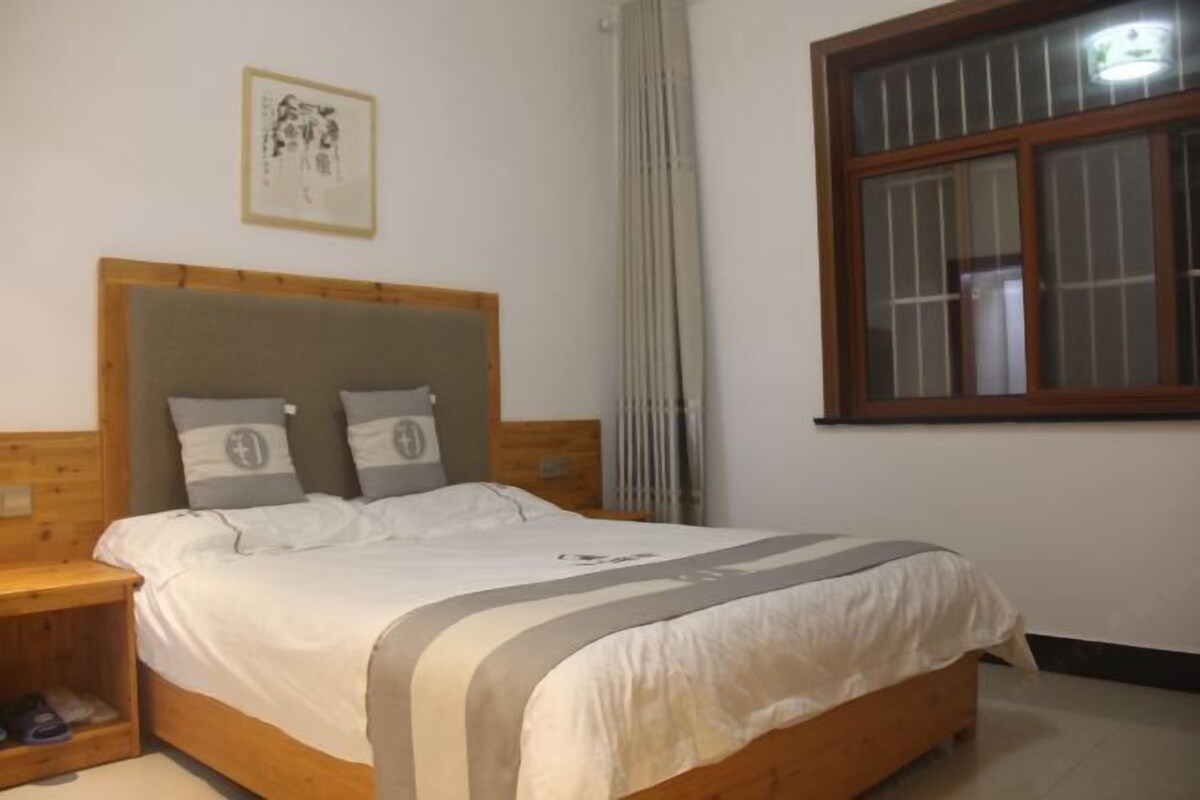 A bedroom with a wooden headboard, a bed with white bedding, a nightstand, a window with blinds, and a framed picture on the wall.