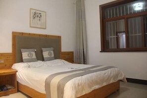 A bedroom with a wooden headboard, a bed with white bedding, a nightstand, a window with blinds, and a framed picture on the wall. - Moutain Jiuhua Jingxin Hotel (Qingyang)
