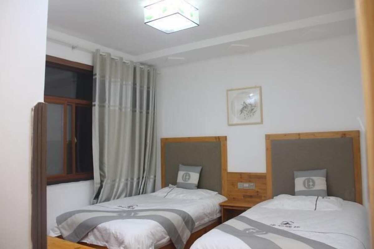 A bedroom with two beds, a window with curtains, a framed picture on the wall, and a ceiling light.