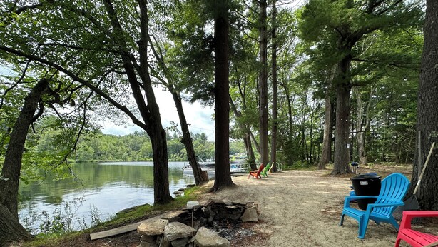 Un camping con una fogata, sillas y vista a un lago rodeado de árboles.