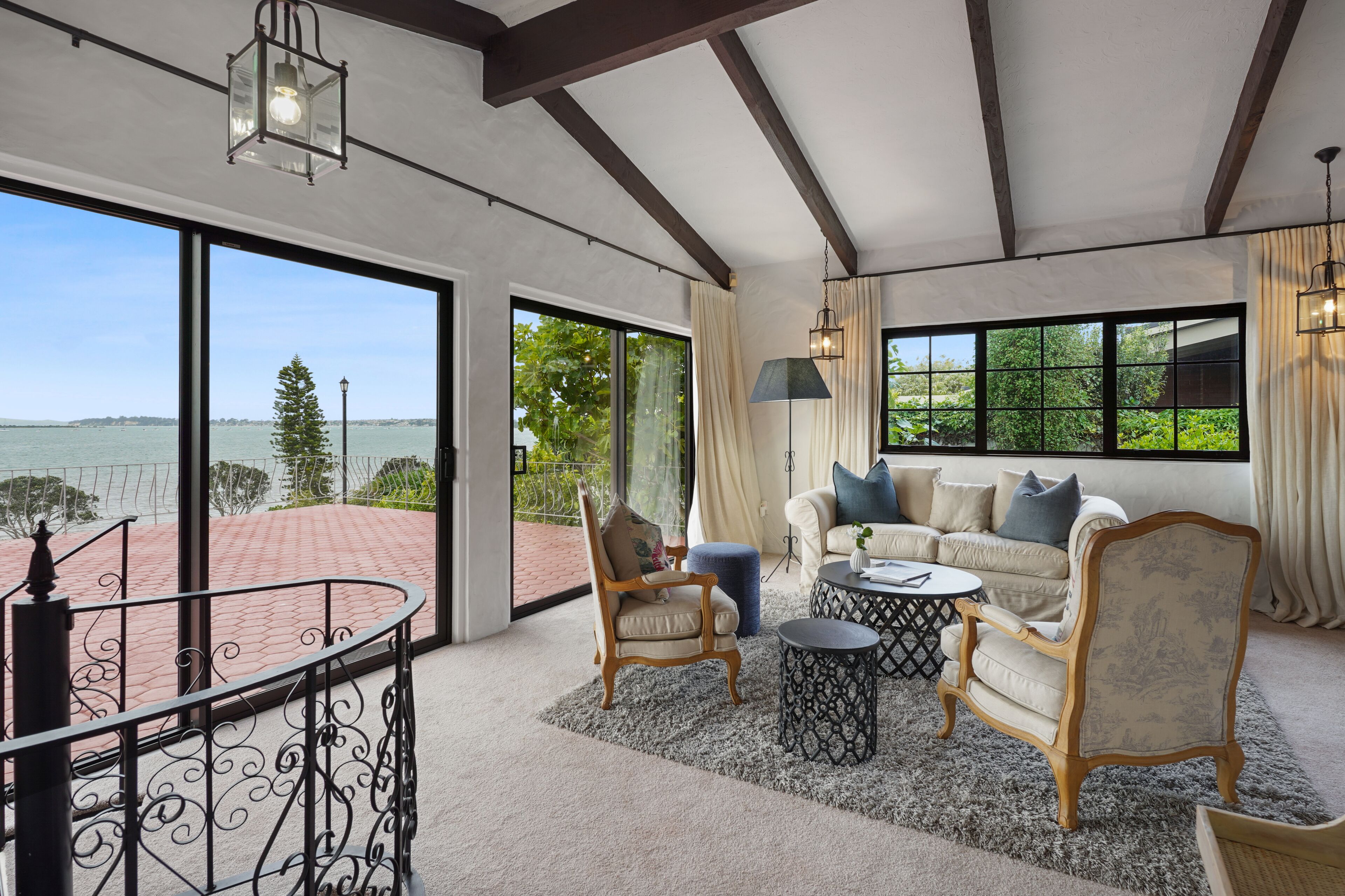 A spacious living room with a large window offering a view of the water, a balcony with a railing, and a cozy seating area with a sofa and chairs.