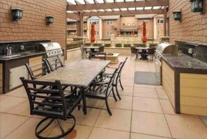 Een terras buiten met een eettafel en stoelen, een grillplaats en een uitzicht op het gebouw op de achtergrond.
