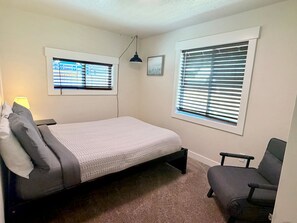 Un dormitorio con una cama individual, dos ventanas con persianas, un escritorio con lámpara y una silla.