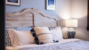 Un lit bien fait, avec une literie à rayures et des coussins décoratifs, une tête de lit en bois et une lampe de chevet agrémentée d’un vase à motifs.