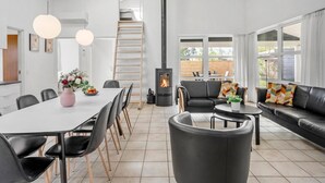 Ein modernes Wohnzimmer mit Kamin, einer Essecke mit weißem Tisch und schwarzen Stühlen sowie einer Couch mit bunten Kissen.