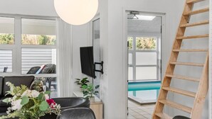 Ein Zimmer mit Esstisch, Stühlen, einem an der Wand montierten Fernseher, einer Holzleiter und Blick auf einen Außenpool.