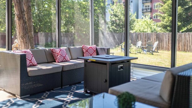 En solbelyst patio med en fletsofa, et lille bord og udsigt over et boligområde.