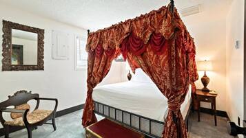 A four-poster bed with ornate red curtains, a wooden chair with a cushion, a bedside table with a lamp, and a vintage suitcase.