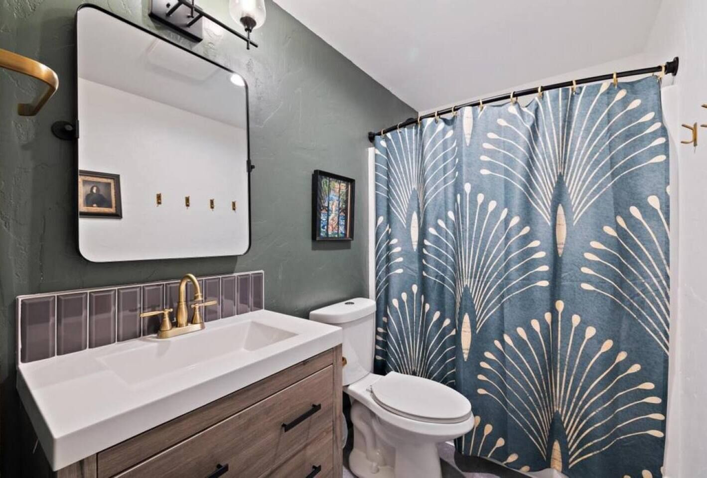 A bathroom with a white sink, gold faucet, and a wooden vanity. There is a white toilet and a shower curtain with a leaf pattern.