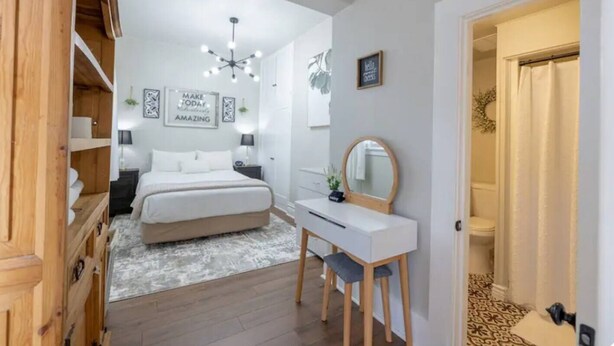 A bedroom with a bed, bedside table, and a desk with a mirror. There is a wall with framed inspirational quotes and a ceiling light fixture. - The Flat on 5th-walk to dt! (Coeur d'Alene)