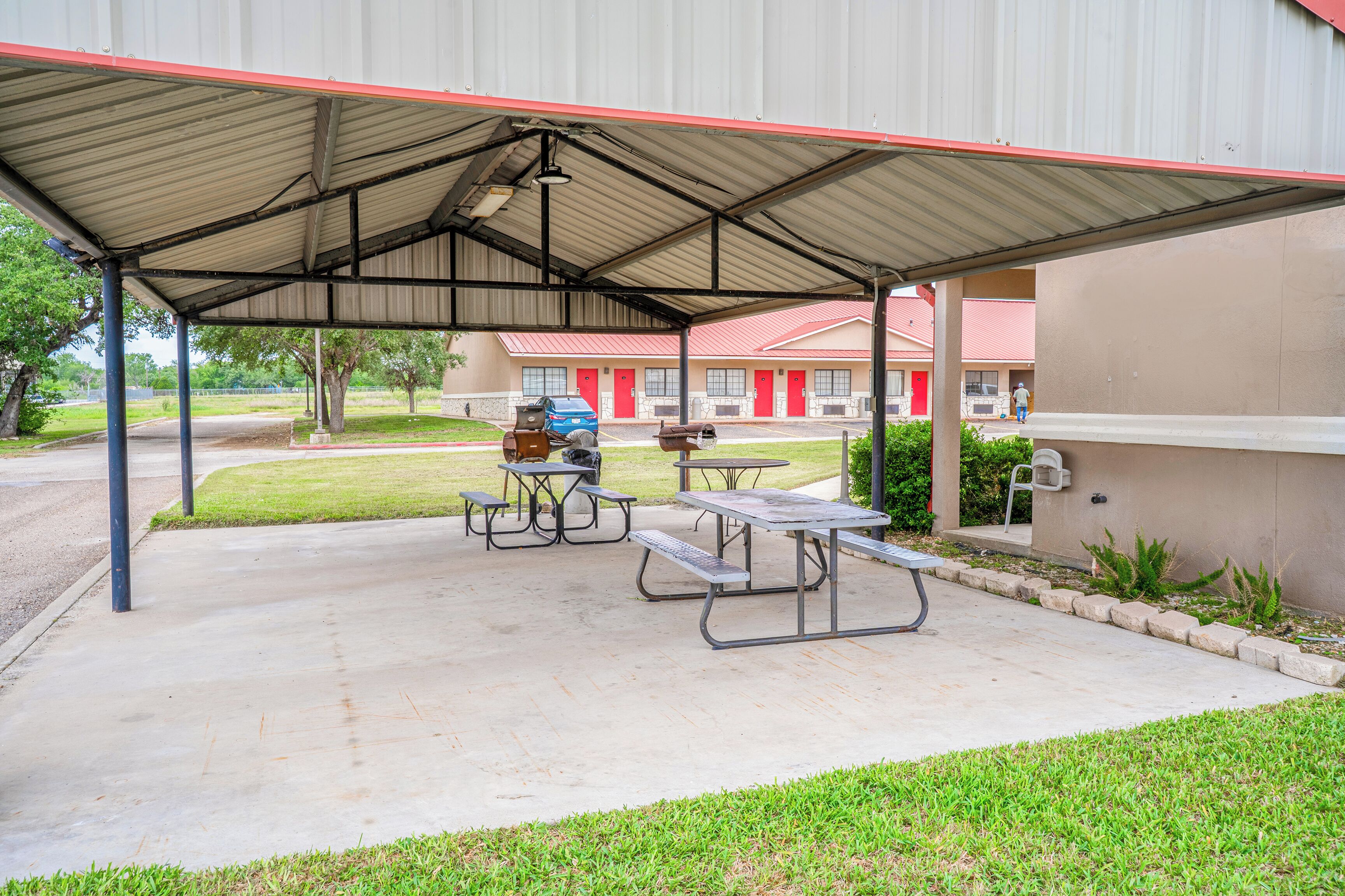 A covered picnic area with tables and benches, a blue car, and a building with red doors in the background.
