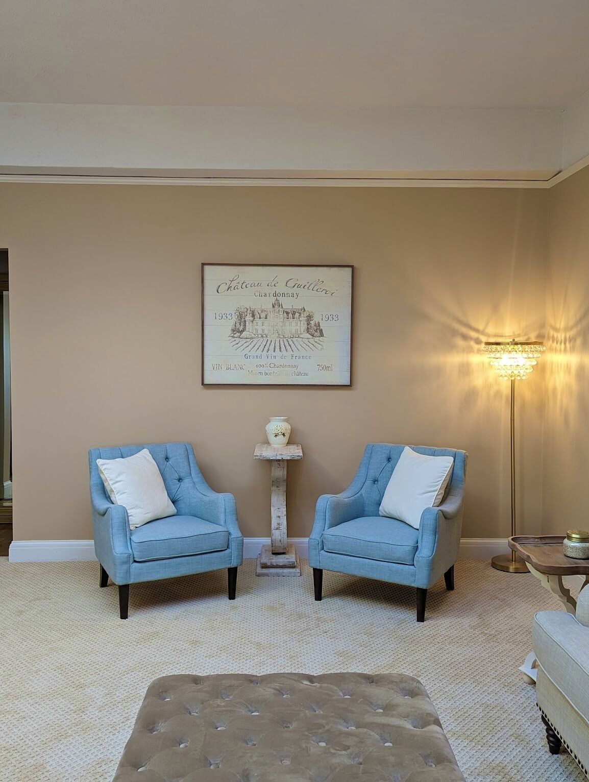 Un salon avec deux fauteuils bleus, une moquette beige, un pouf capitonné, une étiquette de vin encadrée et un lampadaire.