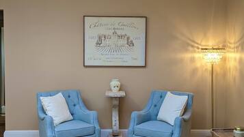 Un salon avec deux fauteuils bleus, une moquette beige, un pouf capitonné, une étiquette de vin encadrée et un lampadaire.
