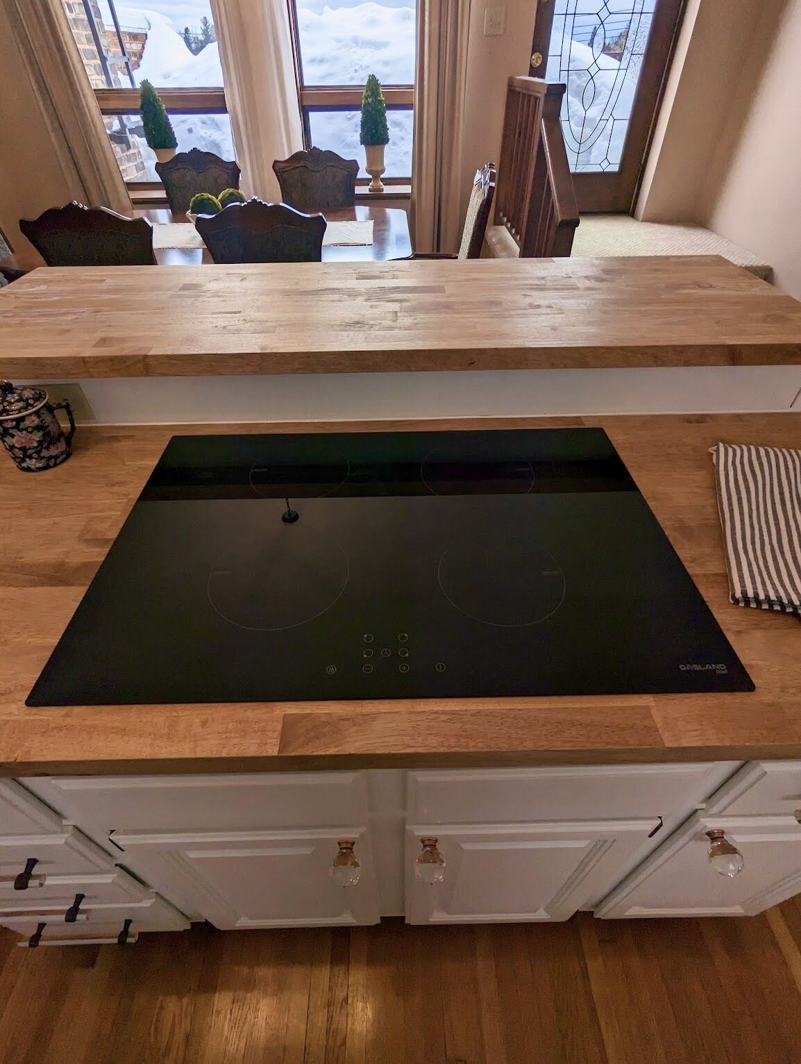 Une cuisine dotée d’une plaque de cuisson à induction noire, de plans de travail en bois, de meubles blancs et offrant une vue sur un paysage enneigé grâce à de grandes fenêtres.
