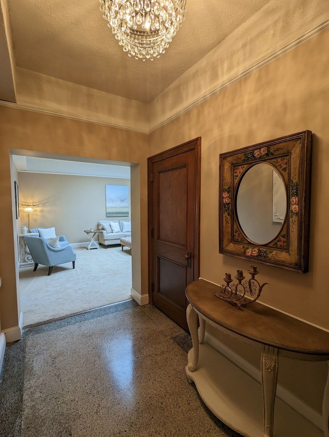 A hallway with a chandelier, a wooden door, a mirror with floral frame, and a seating area with a sofa and armchairs.