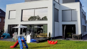 Een modern huis met twee verdiepingen, grote ramen, een patio met tuinmeubilair en een glijbaan voor kinderen.