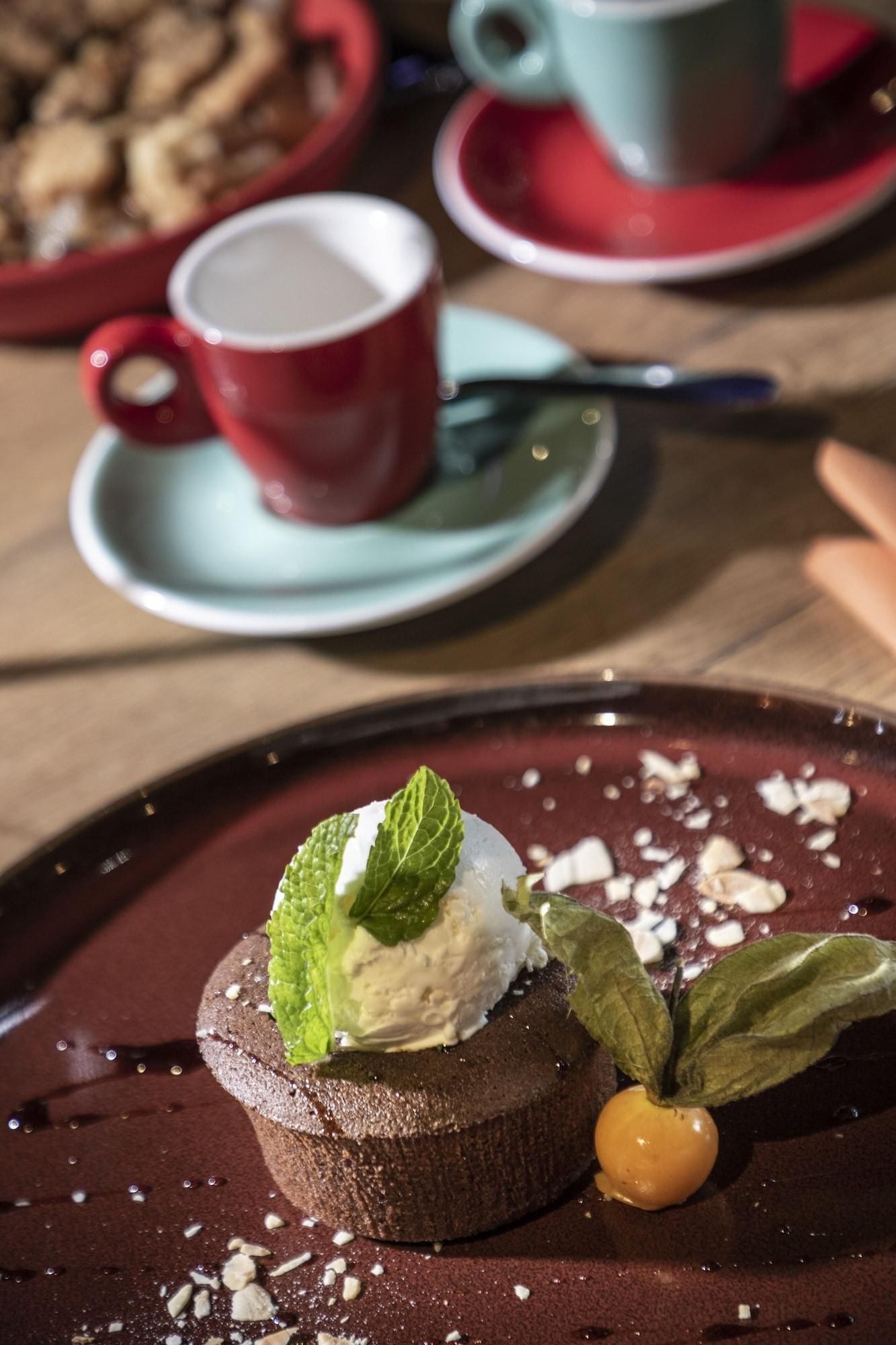 A dessert on a plate with a mint leaf and a small fruit, accompanied by a red cup and saucer in the background.
