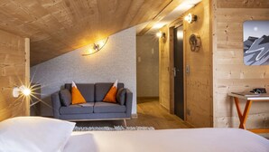 A cozy room with a sloped ceiling, a grey sofa with orange cushions, a bed with white bedding, and a small table with a lamp. - Base Camp Lodge (Bourg-Saint-Maurice)