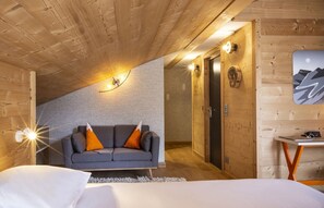 A cozy room with a sloped ceiling, a grey sofa with orange cushions, a bed with white bedding, and a small table with a lamp. - Base Camp Lodge (Bourg-Saint-Maurice)