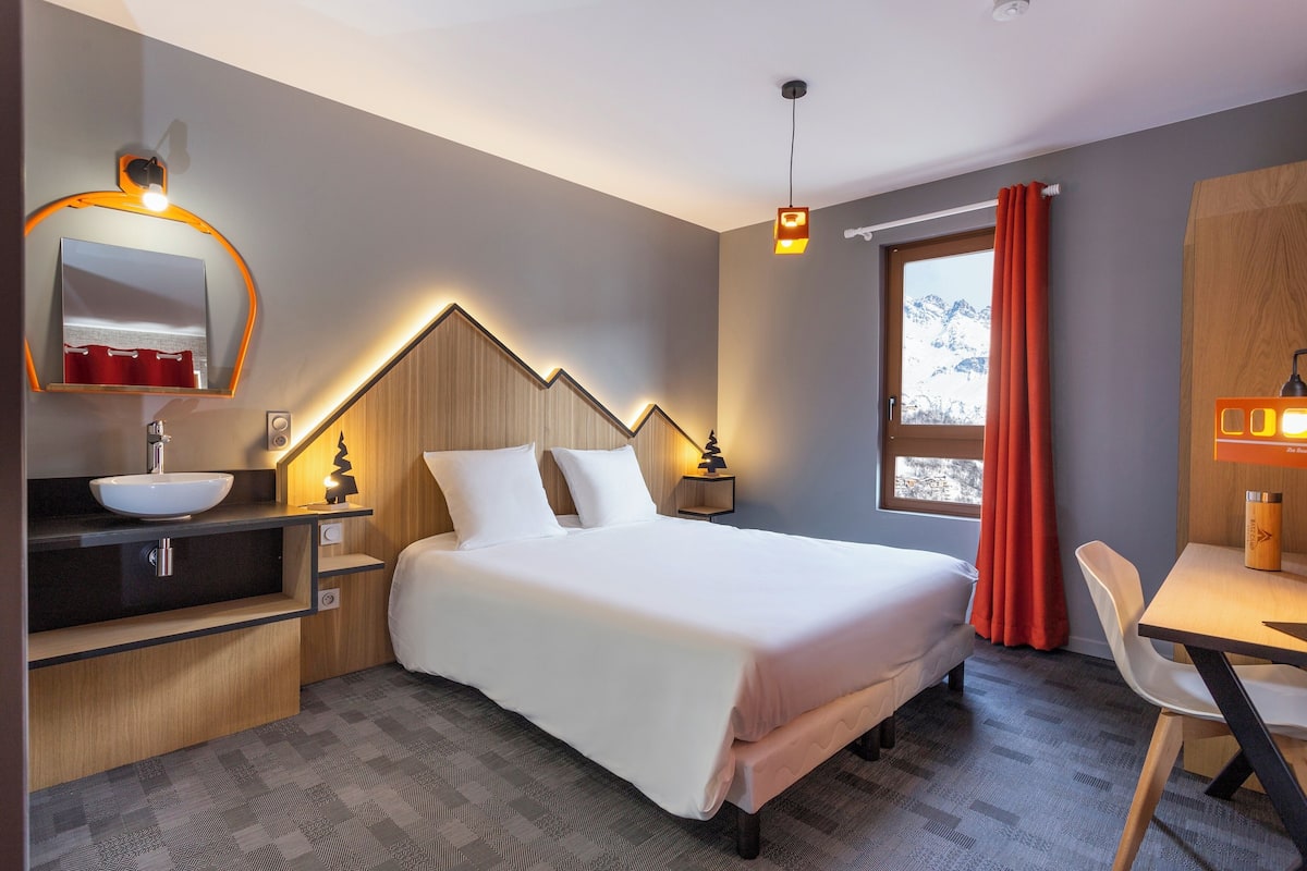 Een hotelkamer met een bed, nachtkastje, bureau, stoel en een badkamer met een wastafel en spiegel.