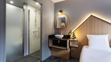 Habitación de hotel con ducha, lavabo, mesita de noche con lámpara y cama.