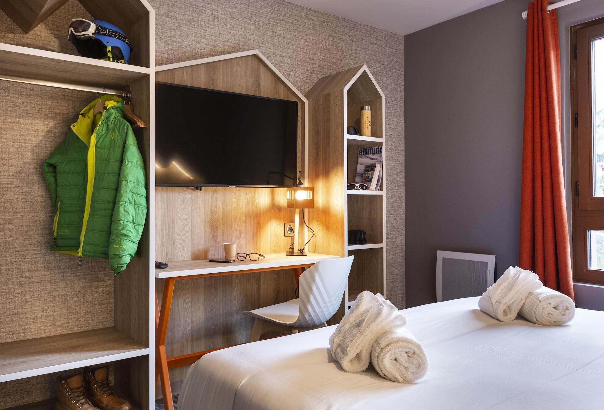 Quarto de hotel com uma cama, uma escrivaninha com televisão, uma cadeira, uma prateleira com um capacete e uma janela com cortinas.