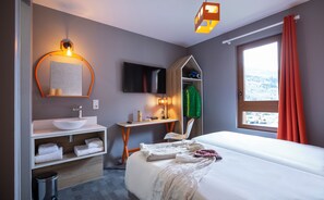 A hotel room with a bed, a bathroom with a sink, a mirror, a TV, and a window with curtains. - Base Camp Lodge (Bourg-Saint-Maurice)