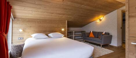 A bedroom with a wooden ceiling, a bed with white bedding, a grey sofa with orange cushions, and a small table.