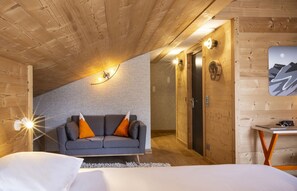 Une pièce chaleureuse avec un plafond en pente, un canapé gris agrémenté de coussins orange, un lit avec une literie blanche et une petite table avec une lampe.