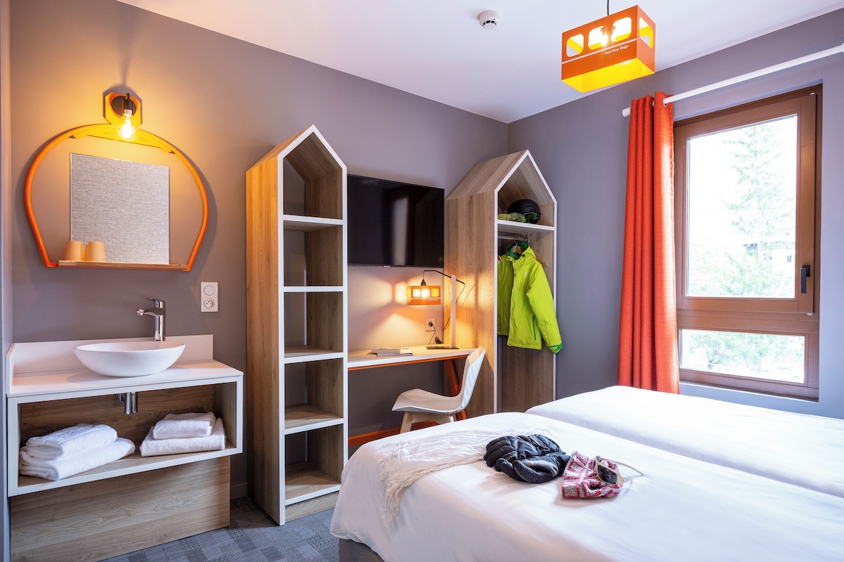 Una habitación de hotel moderna con un lavamanos blanco, una cama grande, un escritorio con una silla y un televisor.