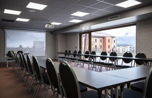 A conference room with a long table, chairs, a projector screen, and large windows with a view of buildings.