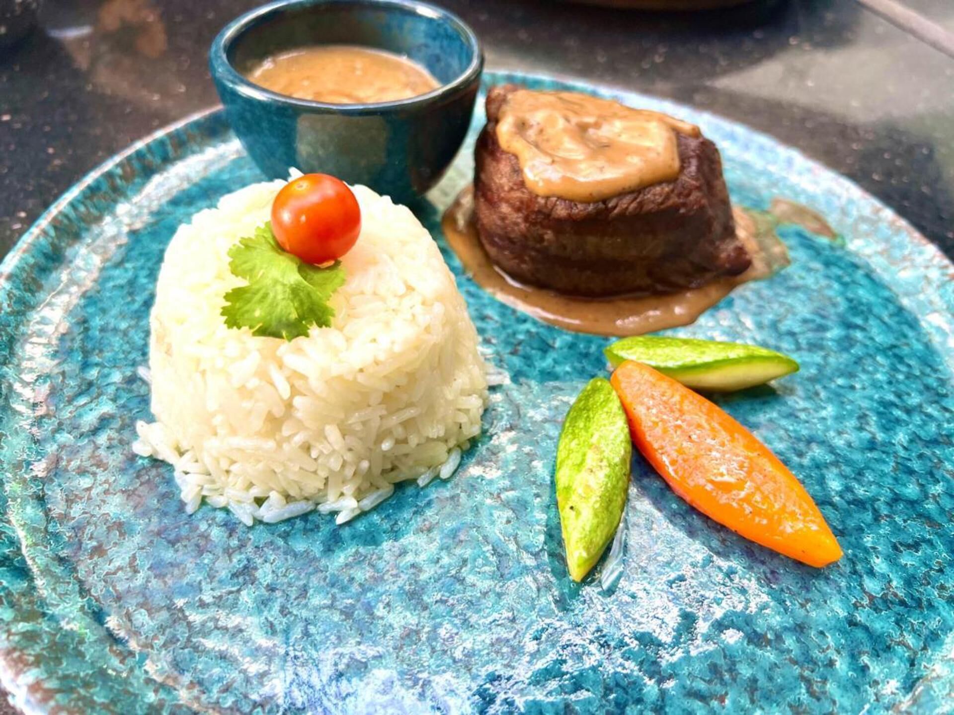 Une assiette de riz blanc garnie d’une tomate cerise et de coriandre, d’un morceau de viande nappé de sauce, ainsi que de tranches de concombre et de carotte.