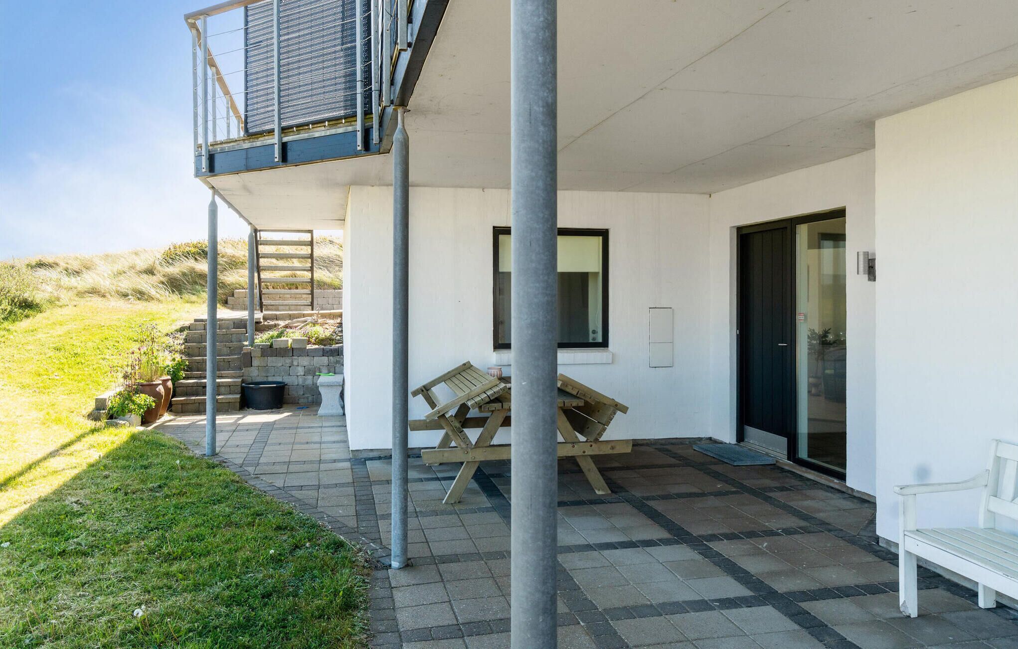 Une terrasse extérieure moderne, avec une table et des chaises en bois, un petit espace jardin agrémenté de plantes et une entrée avec une porte vitrée.