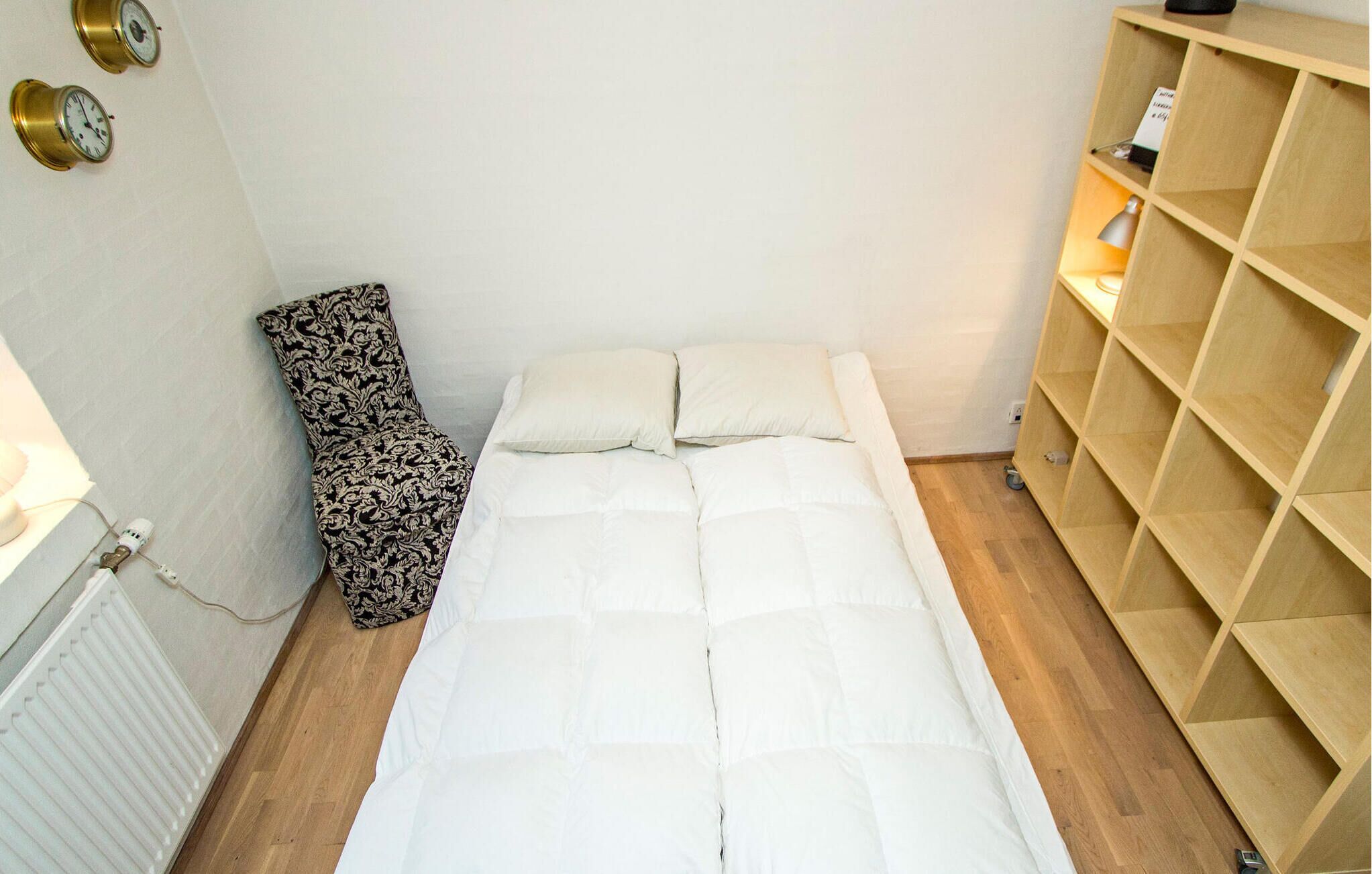 Una camera da letto piccola e ordinata con un letto, cuscini, un termosifone, una sedia con motivo decorativo e una scaffalatura in legno.