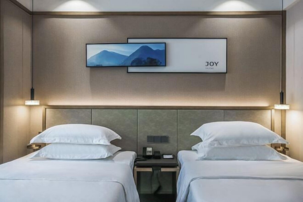Habitación de hotel con dos camas, cabeceros, mesitas de noche y un televisor montado en la pared. Un letrero en la pared indica 'JOY HOTEL'.