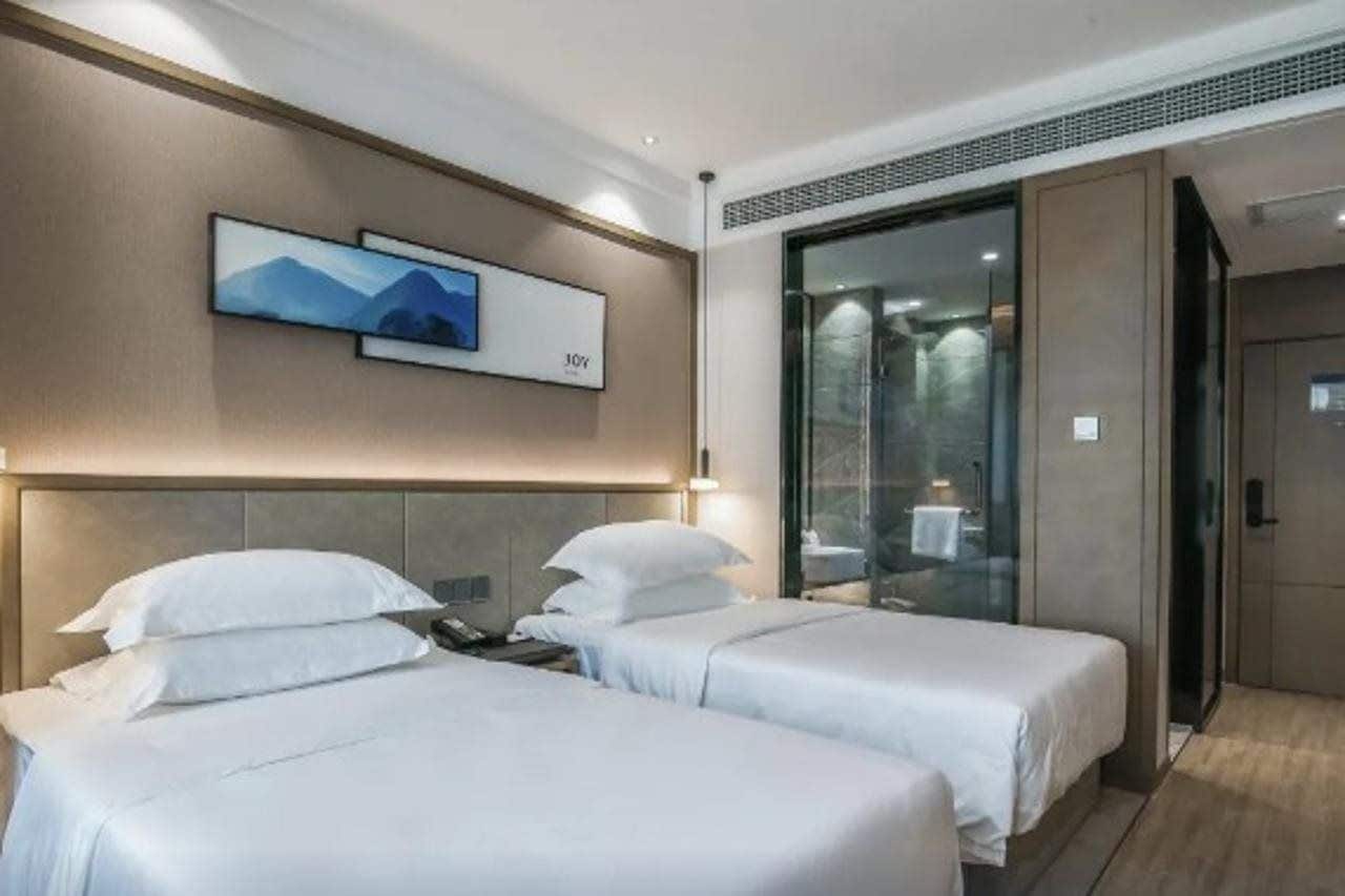Quarto de hotel com duas camas, uma janela grande e uma porta de vidro deslizante que dá para um banheiro.