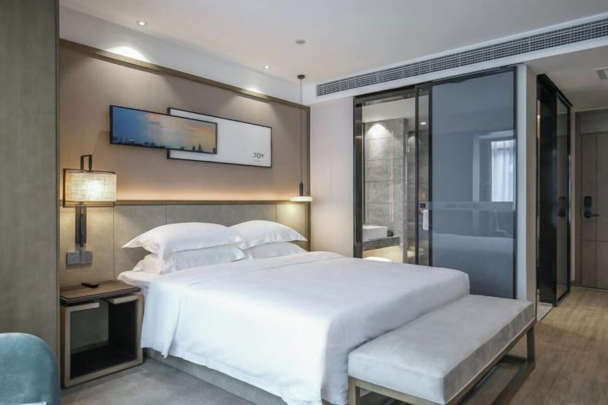 A modern hotel room with a large bed, bedside tables, a wall-mounted lamp, and a sliding glass door to a bathroom.