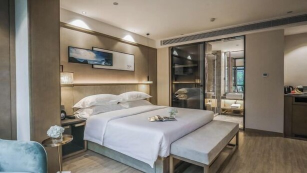 Una habitación de hotel moderna con una cama grande, mesitas de noche, un banco, un vestidor y un baño con un espejo amplio.
