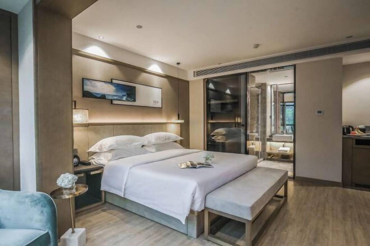 A modern hotel room with a large bed, bedside tables, a bench, a walk-in closet, and a bathroom with a large mirror.