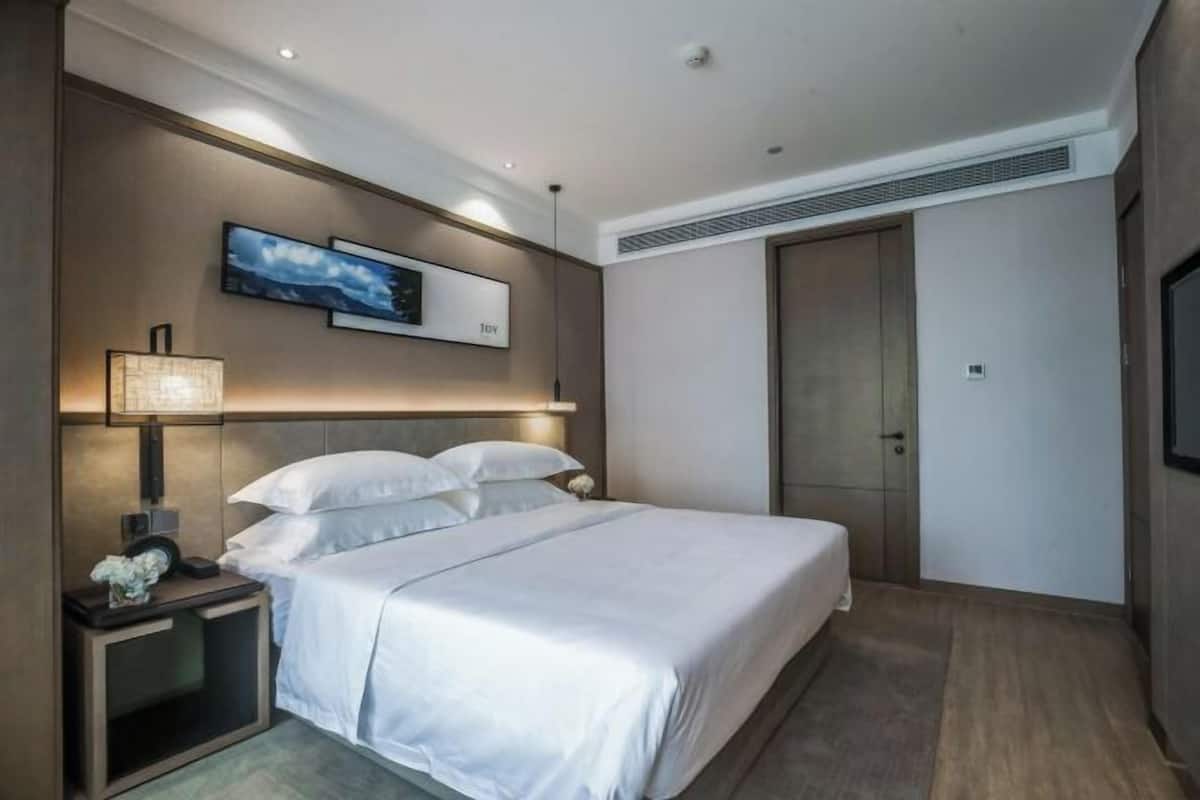 A modern hotel room with a large bed, a bedside table with a lamp, a TV mounted on the wall, and a wardrobe.
