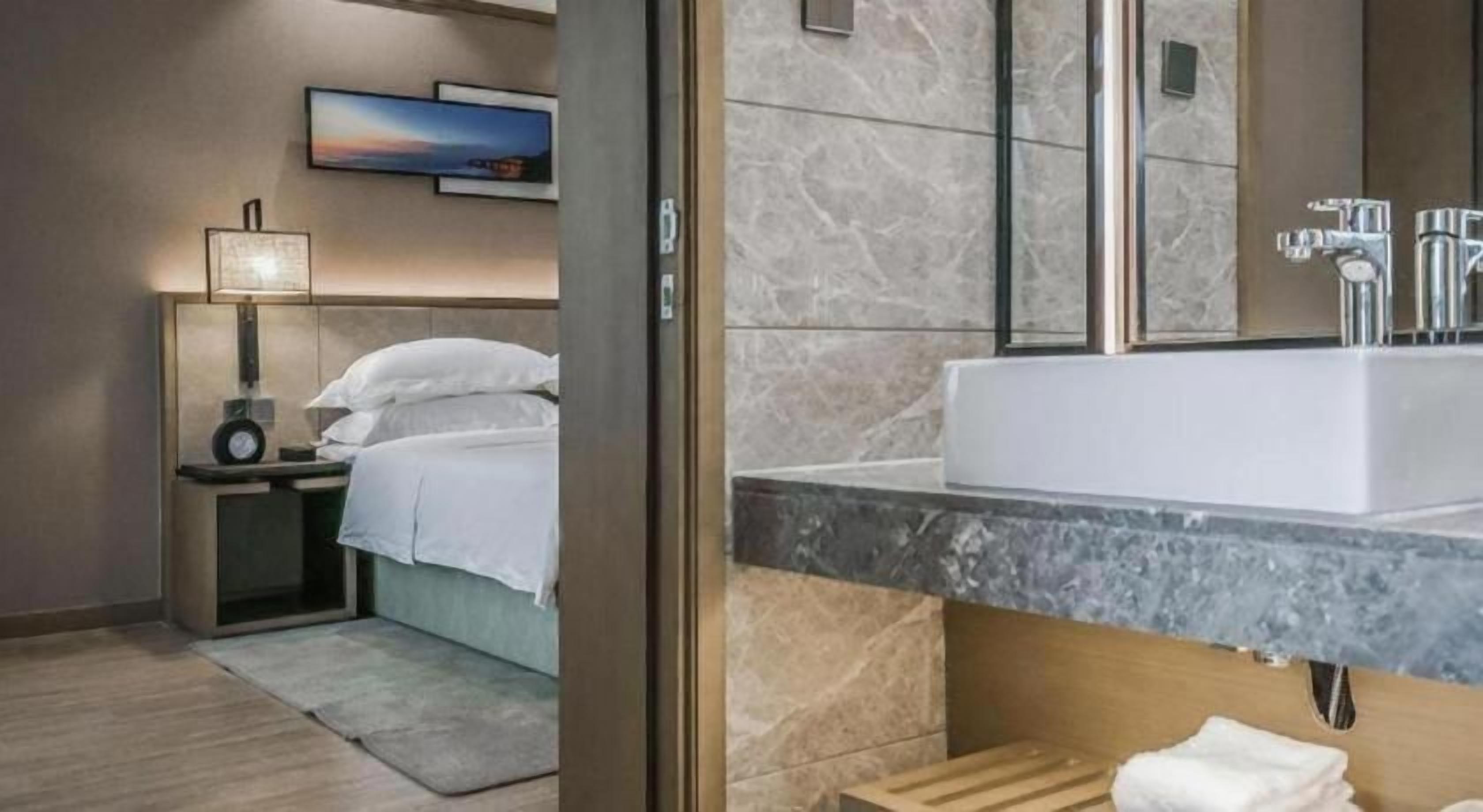 A modern hotel room with a bed, bedside table, lamp, and a large bathroom mirror.
