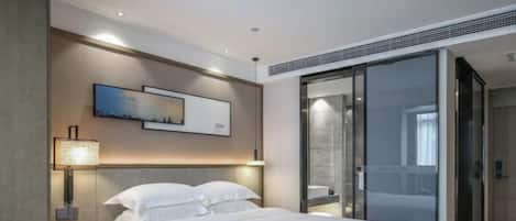 A modern hotel room with a large bed, bedside tables, a wall-mounted lamp, and a sliding glass door to a bathroom.