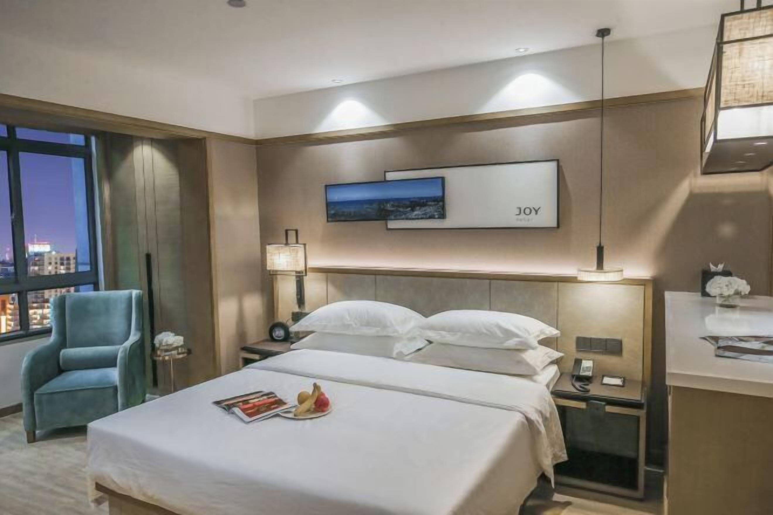 A hotel room with a bed, a nightstand, a lamp, a chair, and a window with a city view.