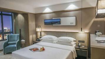 A hotel room with a bed, a nightstand, a lamp, a chair, and a window with a city view.