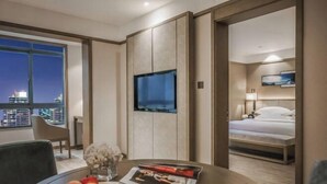 Habitación de hotel con cama, televisor, mesa de comedor con fruta y una revista, y vista nocturna de la ciudad.
