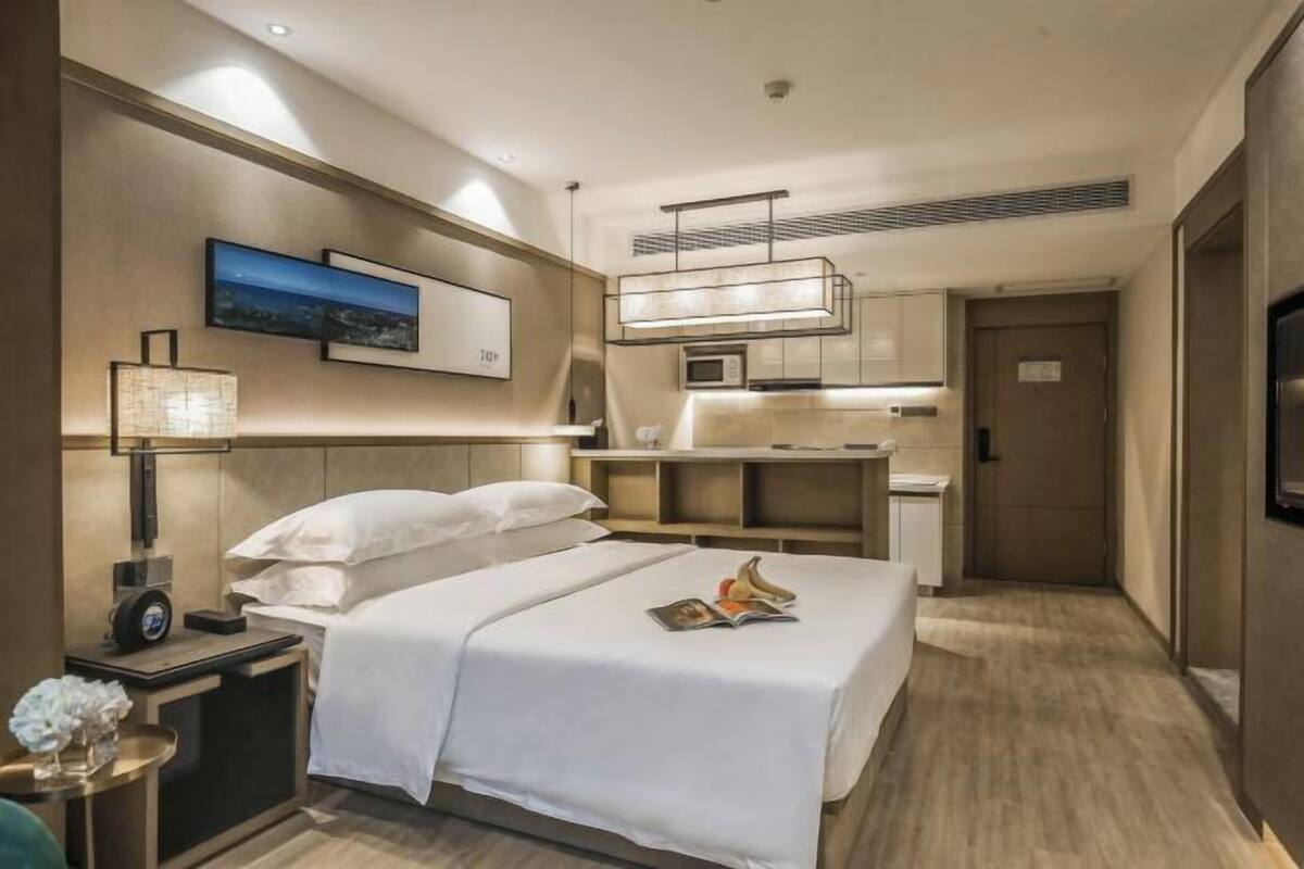 A hotel room with a bed, bedside table, lamp, artwork, and a kitchenette area.
