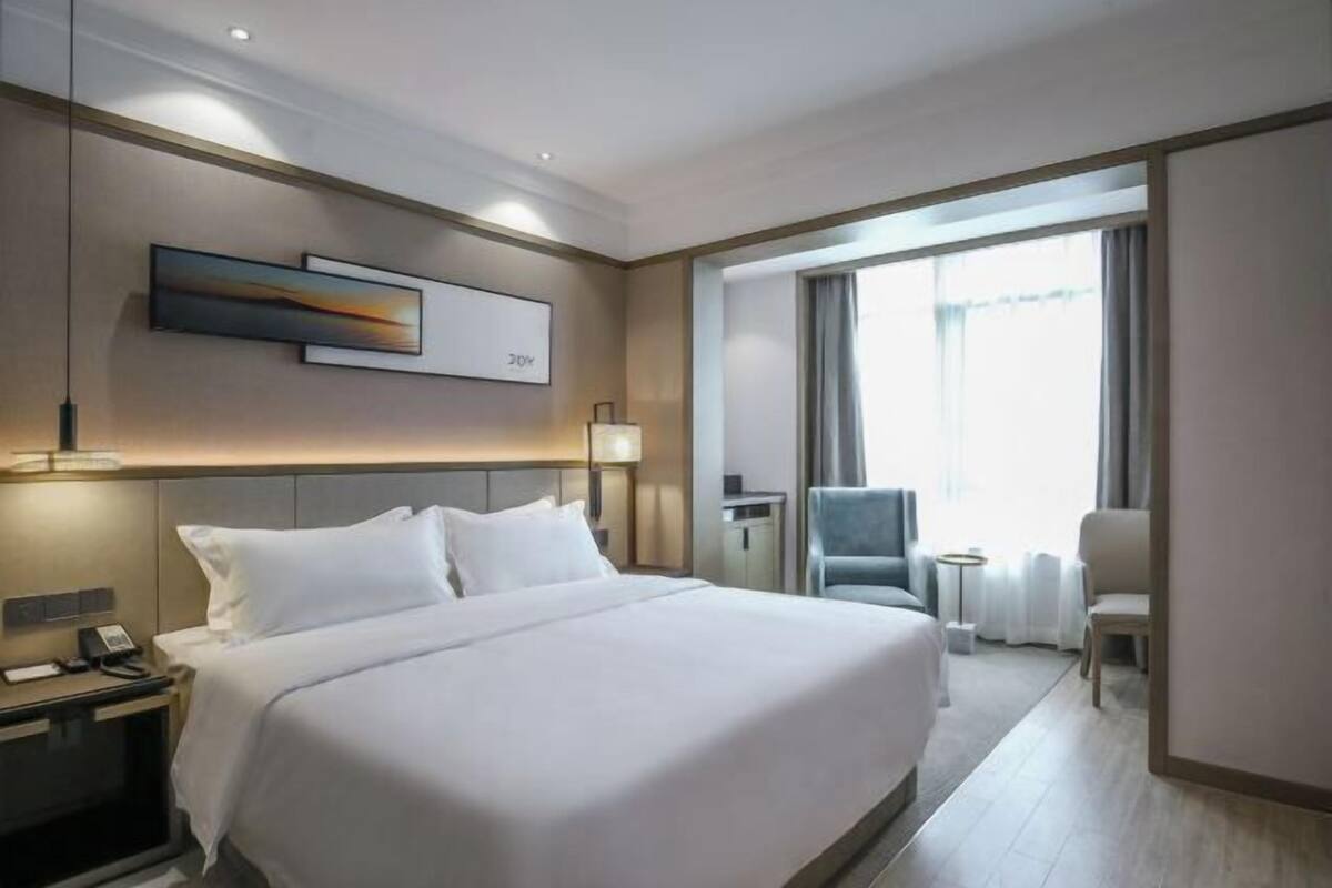 A hotel room with a large bed, a desk with a lamp, a chair, and a view of the outdoors through a large window.