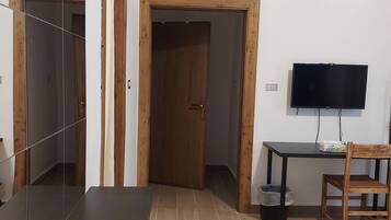 A hotel room with a wooden door, a TV mounted on the wall, a small table with a chair, and a potted plant.
