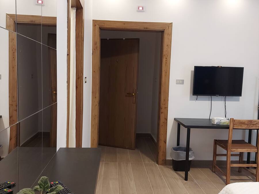 A hotel room with a wooden door, a TV mounted on the wall, a small table with a chair, and a potted plant.